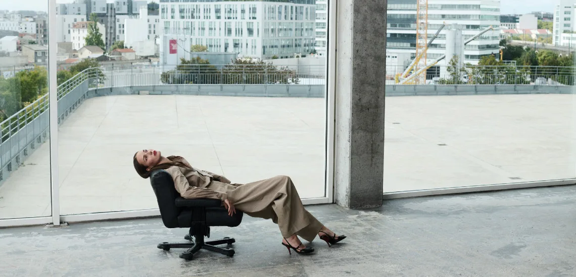 Silhouette en manteau beige et pantalon ample, cheveux détachés, terrasse urbaine avec vue sur la ville
